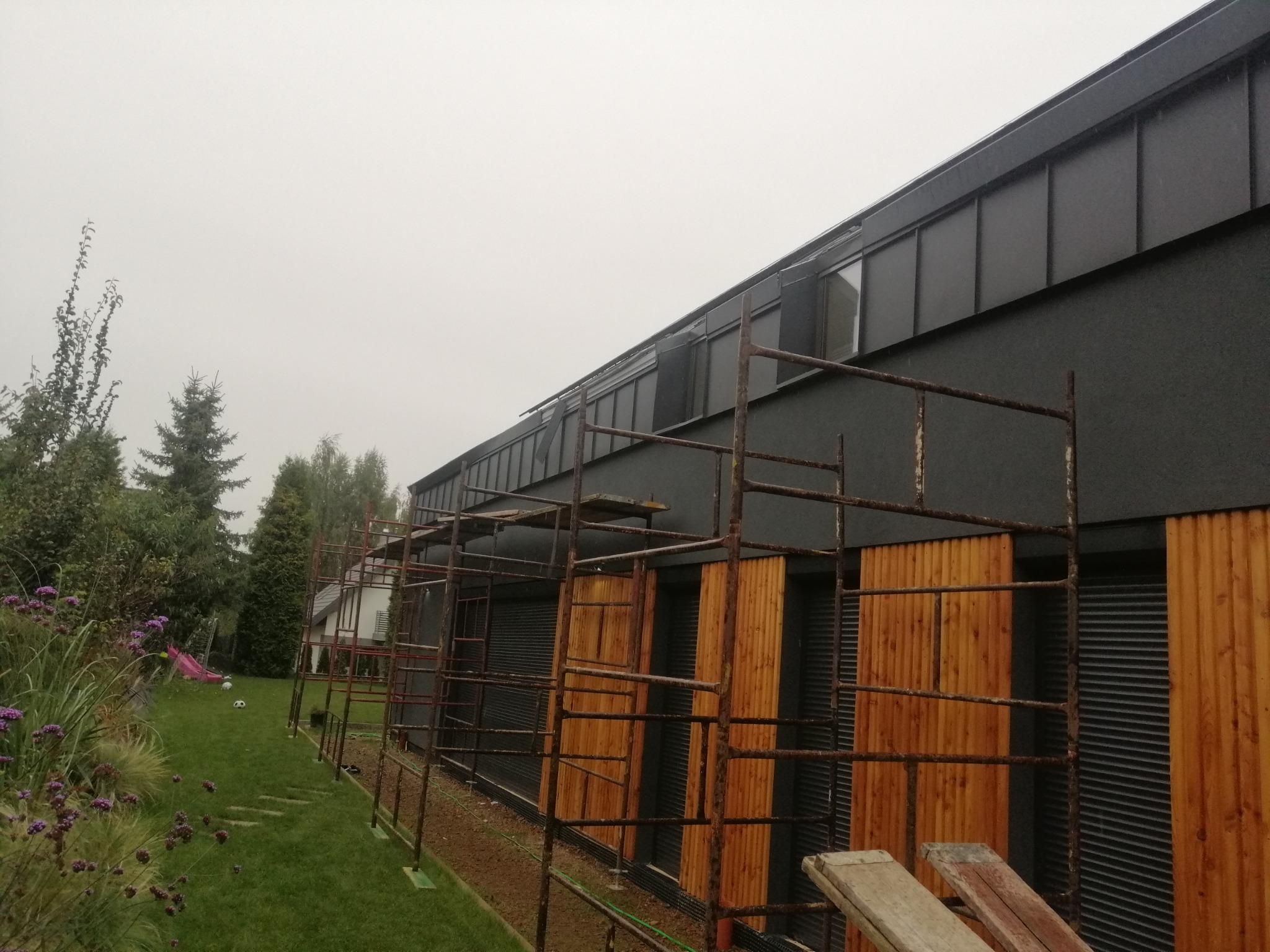 Nowoczesny dom z ciemną elewacją i metalowym dachem, w trakcie prac na wysokości z użyciem rusztowania, widok z ogrodu z elementami zieleni.