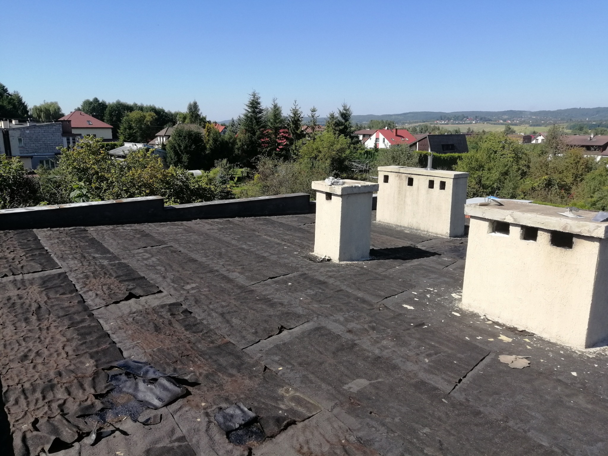 Uszkodzona papa na płaskim dachu z trzema kominami wentylacyjnymi, widoczne pęknięcia i ubytki, w tle zabudowa jednorodzinna i zieleń.