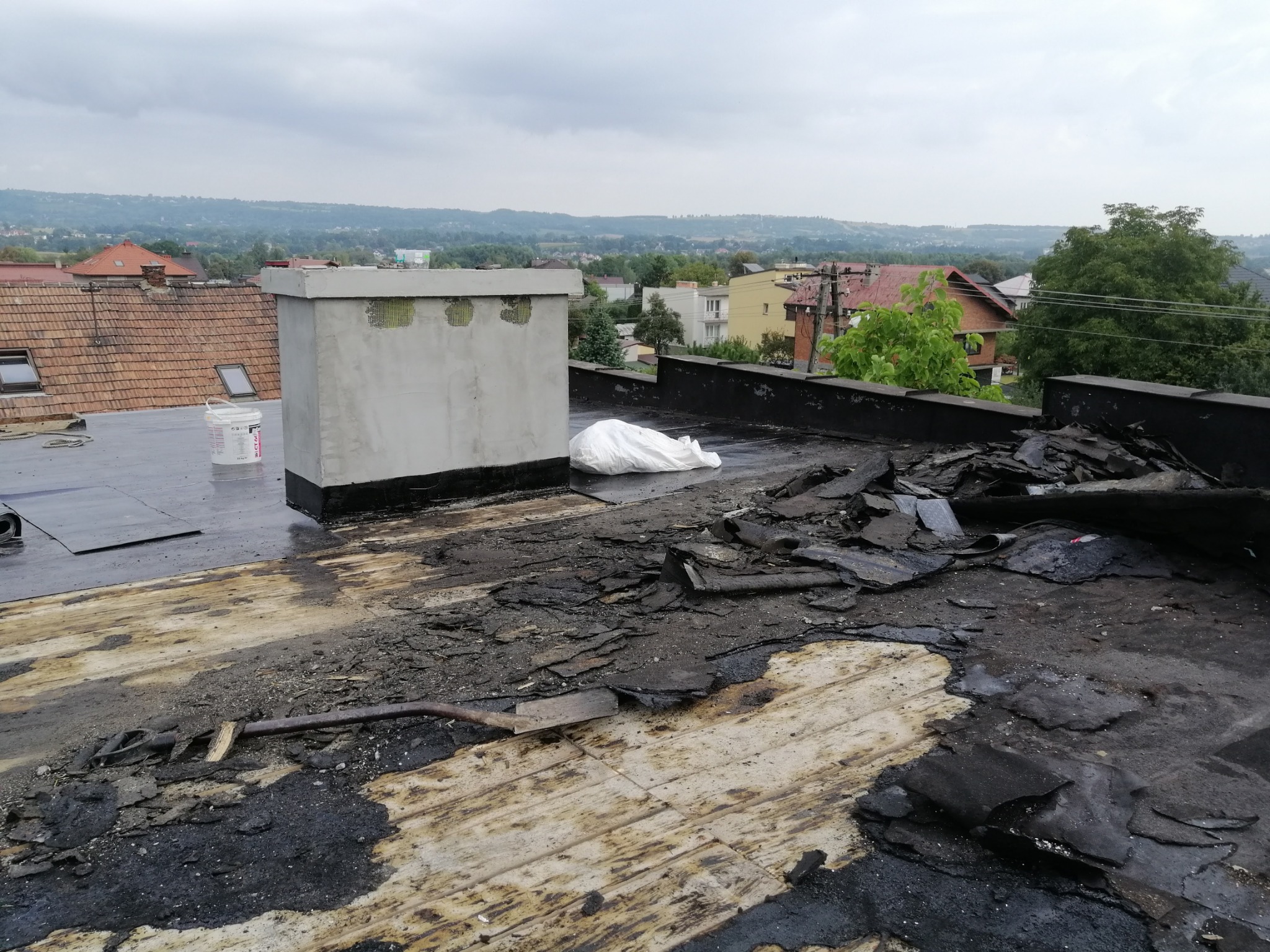 Demontaż starego pokrycia dachowego z papy na płaskim dachu, widoczny fragment komina, wiadro z materiałem budowlanym, narzędzie do usuwania papy oraz widok na okoliczne budynki.