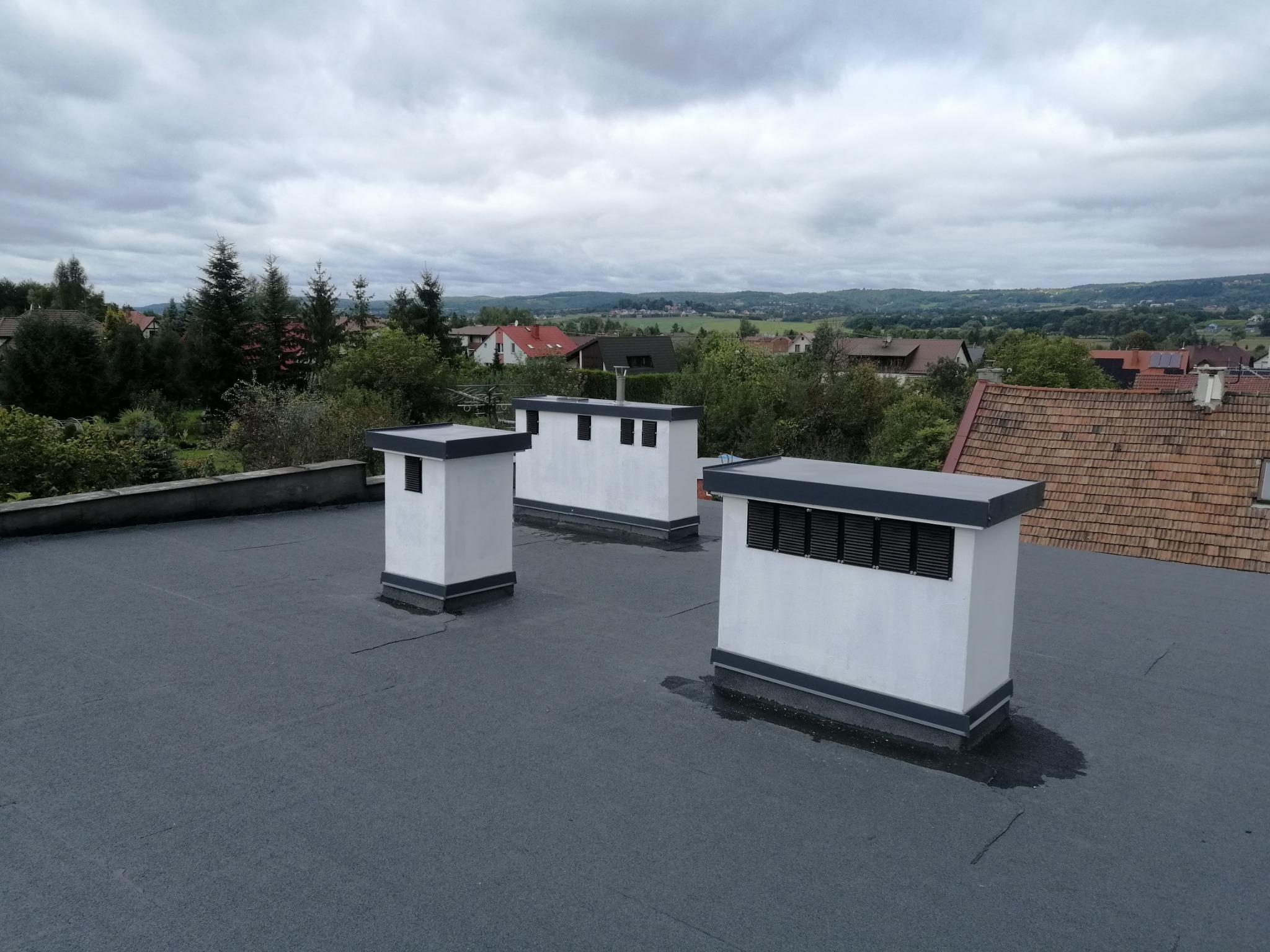 Widok na płaski dach pokryty papą termozgrzewalną z trzema kominami wentylacyjnymi na tle zabudowy jednorodzinnej i pochmurnego nieba.
