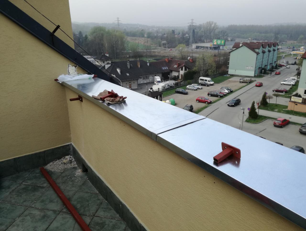 Montaż obróbki blacharskiej na balkonie, widoczne narzędzia i fragment widoku na okolicę.