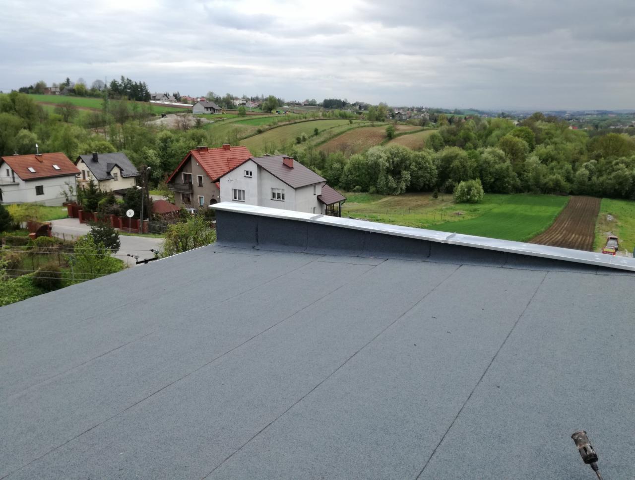 Szary, płaski dach z papy termozgrzewalnej na tle pagórkowatego krajobrazu z zabudowaniami, widok z góry, pochmurne niebo.