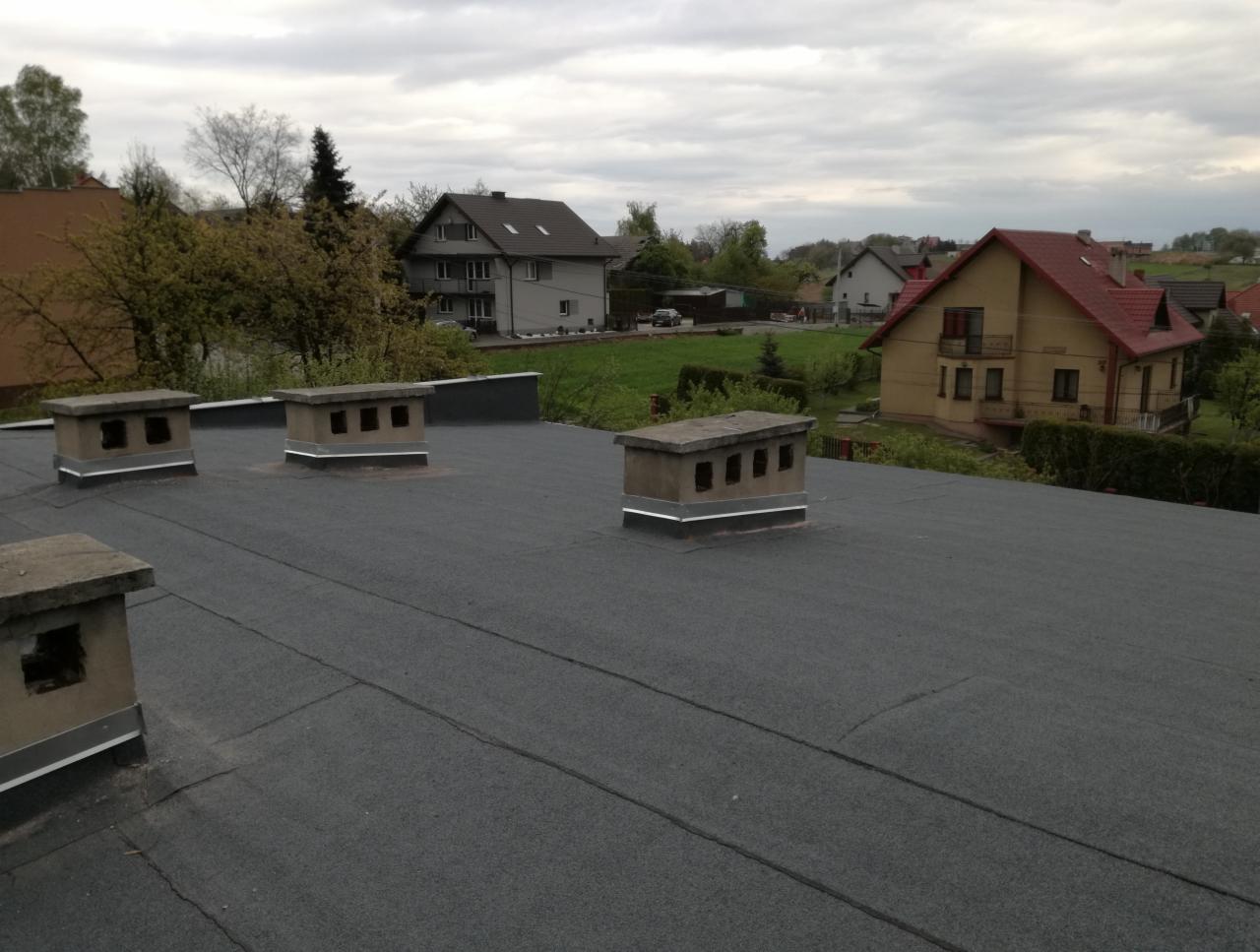 Widok z góry na czarny, płaski dach pokryty papą z czterema betonowymi kominami wentylacyjnymi na tle zabudowy wiejskiej w pochmurny dzień.
