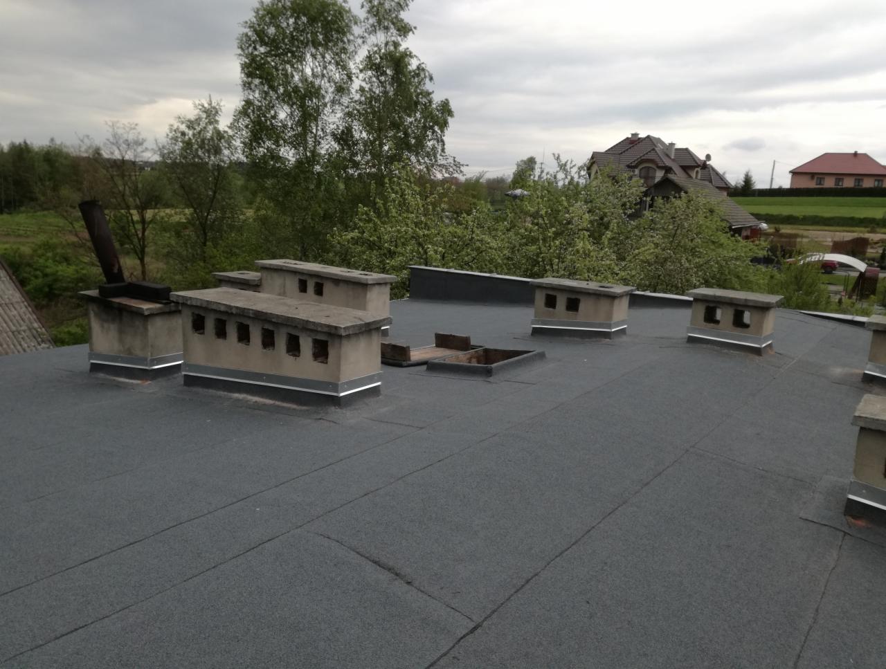 Szary dach pokryty papą termozgrzewalną z wystającymi kominami wentylacyjnymi i kominem dymowym w tle drzewa i zabudowa jednorodzinna.