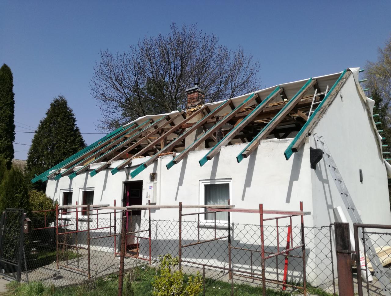 Dom jednorodzinny w trakcie prac dekarskich: odsłonięta konstrukcja więźby dachowej z zielonymi łatami, częściowo zabezpieczona folią, drabina oparta o ścianę, rusztowanie w tle.