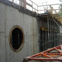 Żelbetowa ściana fundamentowa z okrągłym otworem, szalunki i podpory widoczne w tle, data wykonania zdjęcia w prawym dolnym rogu.