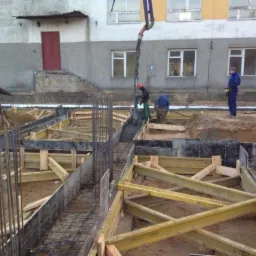 Wylewanie betonu do szalunków fundamentowych zbrojonych stalą, widoczni pracownicy budowlani oraz pompa do betonu na tle budynku.