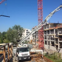 Budowa wielopiętrowego budynku z użyciem dźwigu i pompy do betonu; widoczne zbrojenia i szalunki, w tle zabudowa miejska.