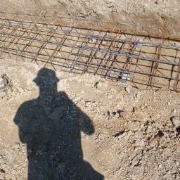Zbrojenie fundamentu w wykopie, widoczny cień osoby w kapeluszu.