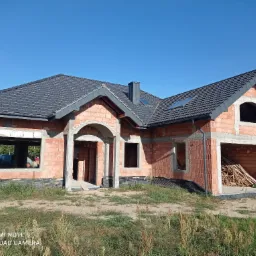 Surowy, jednopiętrowy dom z czerwonej cegły w trakcie budowy, z ciemnoszarą dachówką, widoczne otwory okienne i drzwiowe, stos drewna w garażu, błękitne niebo w tle.