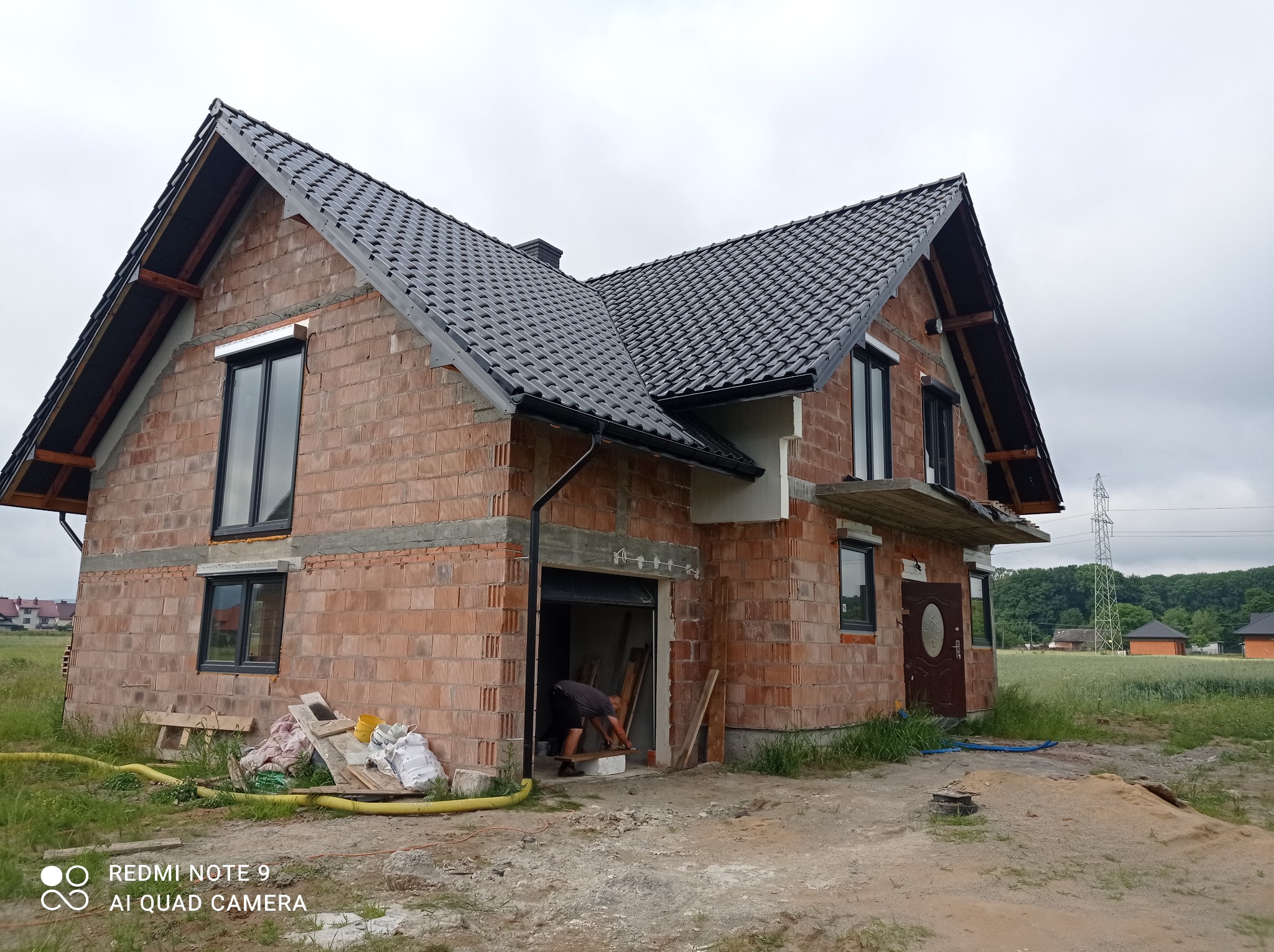 Surowy dom murowany z ciemną dachówką w trakcie budowy, widoczne cegły, okna z czarnymi ramami, mężczyzna w garażu, niebo pochmurne.