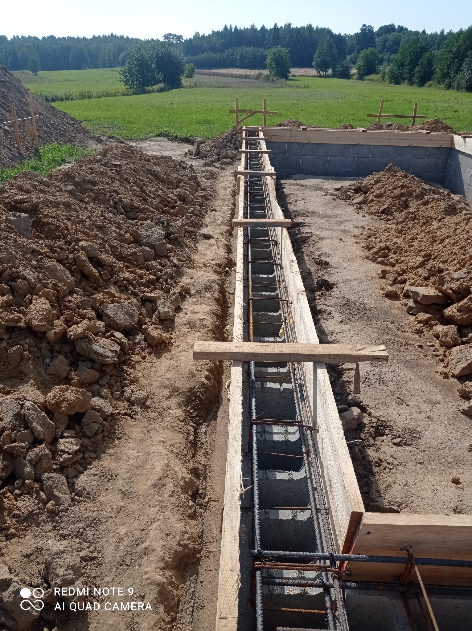 Wykop pod fundamenty z deskowaniem i zbrojeniem stalowym, widok z góry na tle zieleni i drzew.