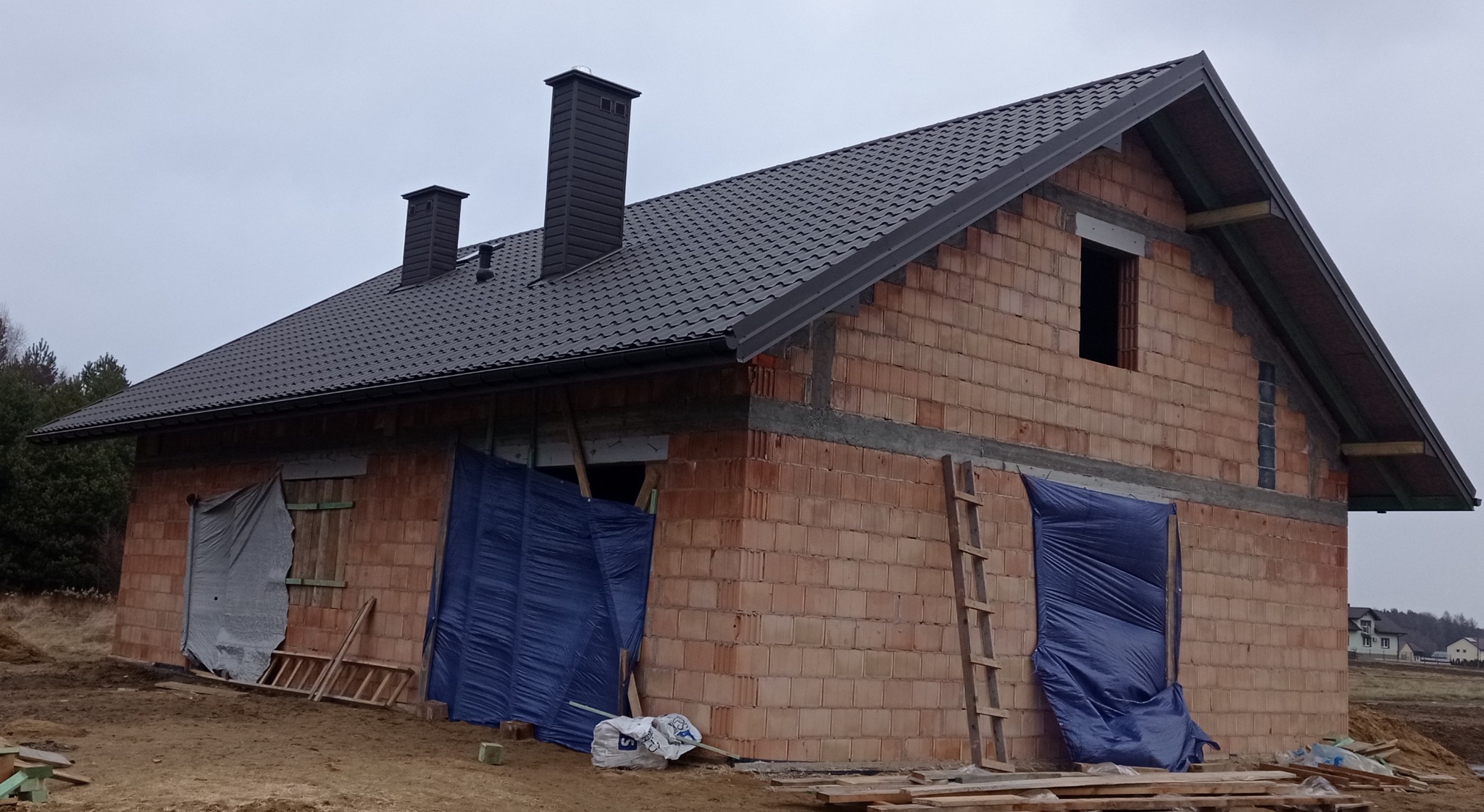Surowy budynek z czerwonej cegły w trakcie budowy, z czarnym dachem i dwoma kominami, zabezpieczony plandekami, z drewnianą drabiną opartą o ścianę.