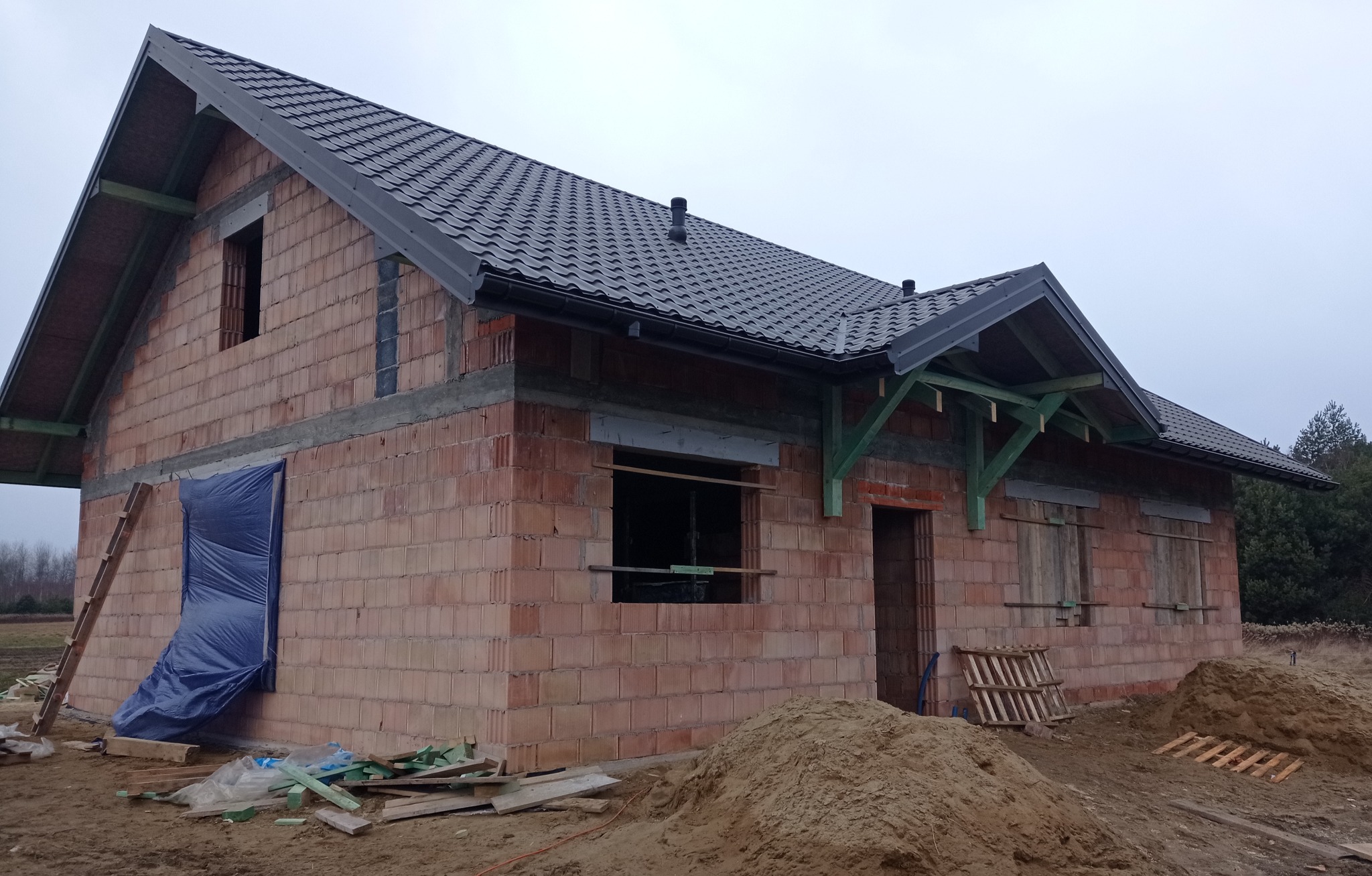Surowy stan domu z czerwonej cegły, z ciemną dachówką, zabezpieczone otwory okienne, drabina oparta o ścianę, hałda piachu przed budynkiem, pochmurne niebo.