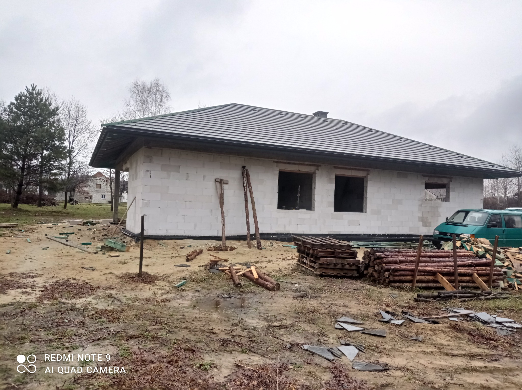 Niedokończony dom jednorodzinny z białego bloczka, z zamontowanym ciemnoszarym dachem, widoczne drewniane stemple, palety i stosy drewna na błotnistym terenie, zielony samochód dostawczy w tle.