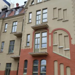 Odnowiona elewacja kamienicy z elementami z czerwonej cegły i jasnobeżowego tynku. Widoczne balkony i okna z zielonymi ramami.