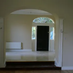 Jasne wnętrze z łukowatym przejściem do przedpokoju z czarnymi drzwiami wejściowymi i grzejnikiem. Ściany w odcieniach beżu, podłoga wyłożona błyszczącymi płytkami.