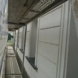 Elewacja budynku po tynkowaniu, widok z rusztowania. Jasna fasada z poziomymi podziałami. Fragment siatki ochronnej po lewej stronie, okna z czarnymi parapetami.