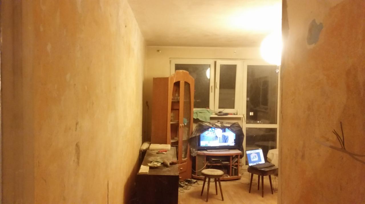 Pokój w trakcie remontu: ściany z usuniętą tapetą, widoczne ślady po kleju. Meble odsunięte, telewizor włączony. Widoczne przygotowania do malowania ścian.