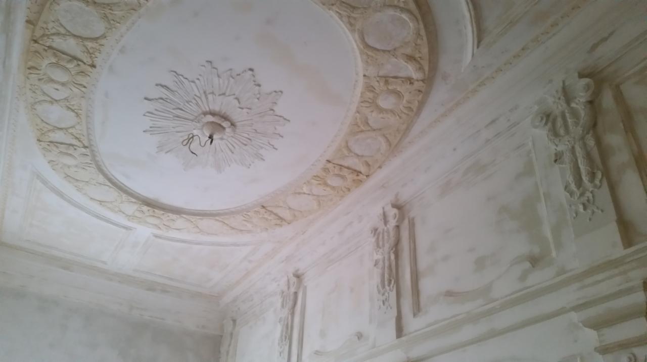 Wnętrze z dekoracyjnymi sztukateriami na ścianach i suficie, w jasnej kolorystyce. Widoczne detale zdobień i delikatne cieniowanie, tworzące klasyczny wygląd.