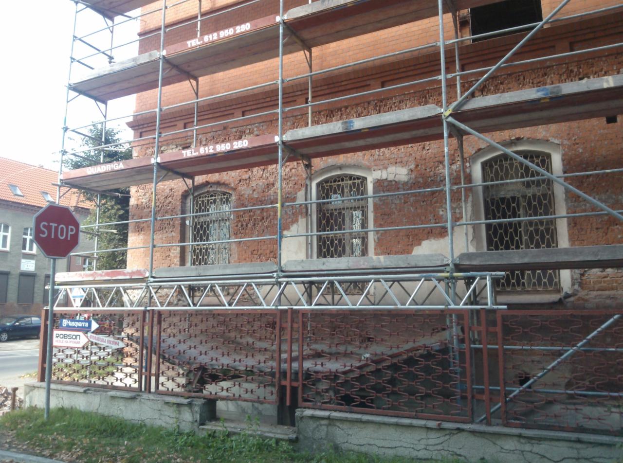 Ceglana elewacja budynku w trakcie renowacji, widoczne rusztowania i fragment ulicy ze znakiem STOP. Okna zabezpieczone kratami.