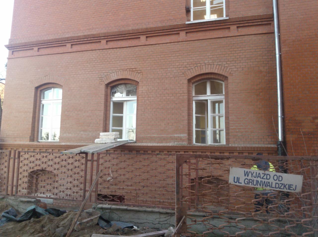 Ceglana elewacja budynku z trzema oknami o łukowatych nadprożach, zabezpieczona metalowym ogrodzeniem z widocznym znakiem informacyjnym.