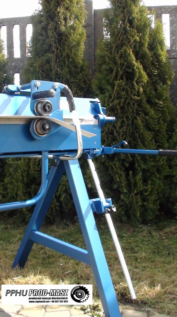 Niebieska zaginarka dekarska PROD-MASZ 3140 0,8 na tle zielonych iglaków i betonowego muru, z widoczną blachą między rolkami.