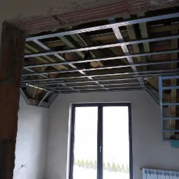 Montaż metalowej konstrukcji sufitu podwieszanego z widocznymi drewnianymi elementami nośnymi, surowy mur przy otworze drzwiowym, okno z widokiem na zewnątrz.