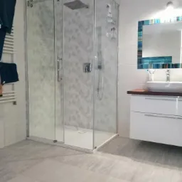 Nowoczesna łazienka z kabiną prysznicową typu walk-in, szare płytki z subtelnym wzorem, biała szafka pod umywalką z drewnianym blatem i lustro z dekoracyjną ramą w odcieniach błękitu.