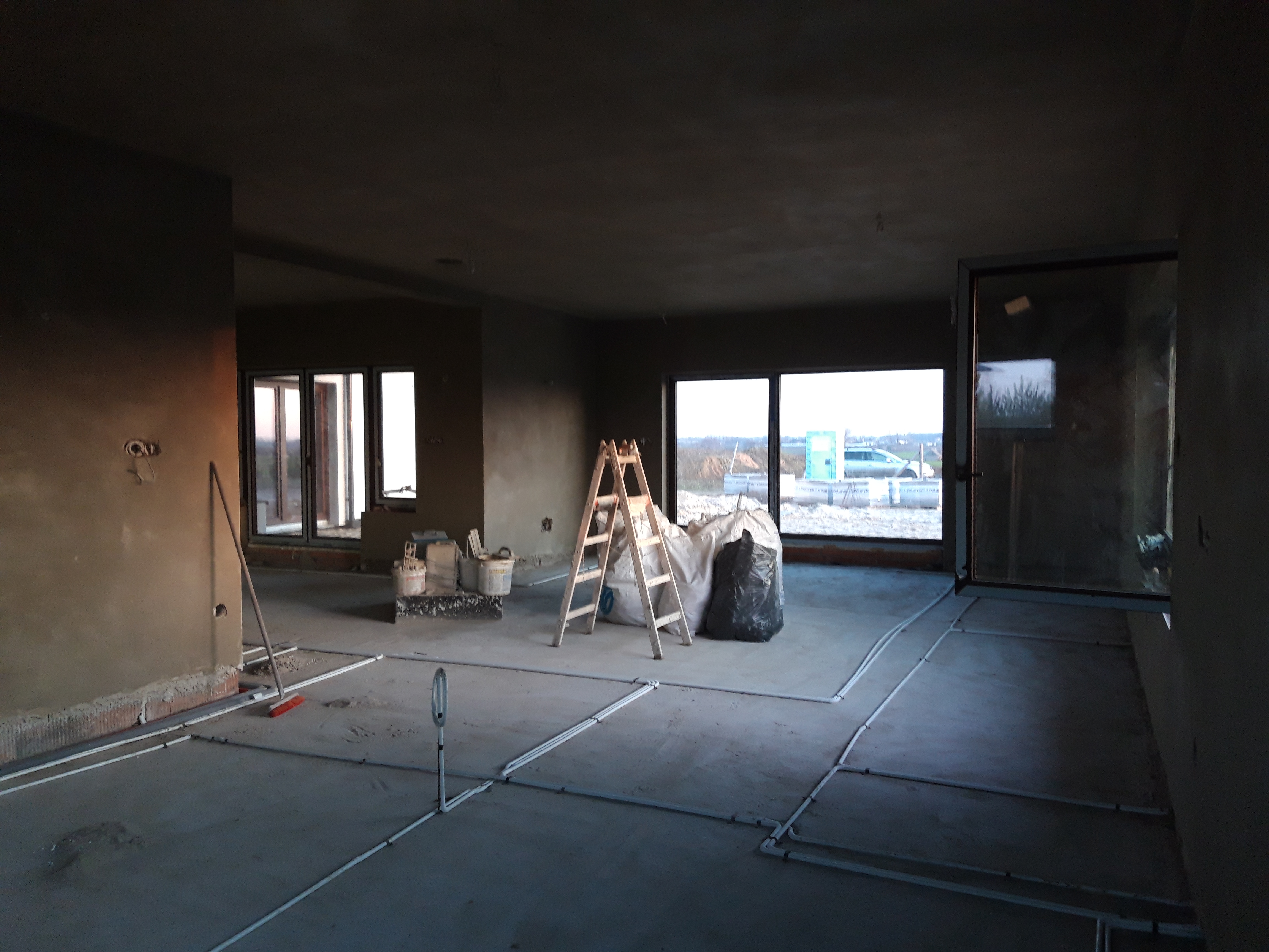 Wnętrze w stanie surowym z nałożonymi tynkami, widoczne okna, drabina, worki z materiałami budowlanymi i instalacja ogrzewania podłogowego przed wylewką.