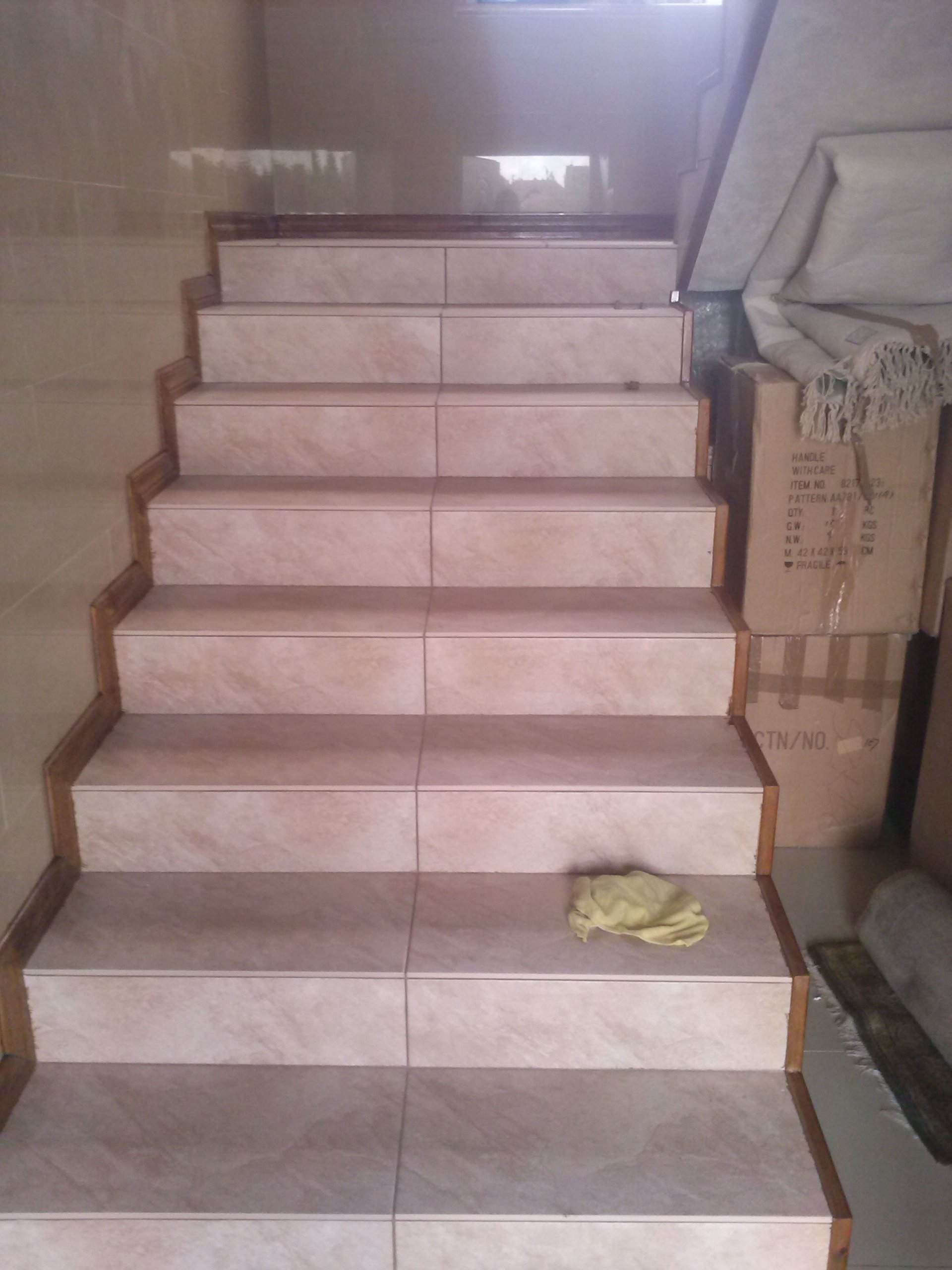 Schody wyłożone jasnymi płytkami ceramicznymi z drewnianymi listwami wykończeniowymi, na jednym ze stopni leży żółta ścierka, a w tle widoczne kartony z tekstem 'HANDLE WITH CARE'.