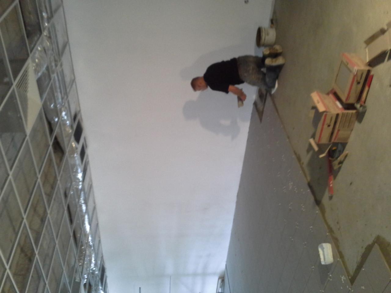 Praca tynkarska: Tynkarz aplikuje gładź na sufit w pomieszczeniu z oknami z luksferów. Na podłodze leżą kartony i narzędzia.