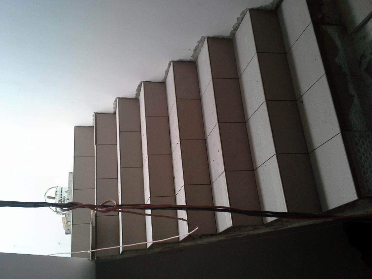 Surowe schody z szarymi płytkami, widok z dołu, z widocznymi przewodami elektrycznymi i niedokończonym wykończeniem sufitu, ukazujące etap prac budowlanych.