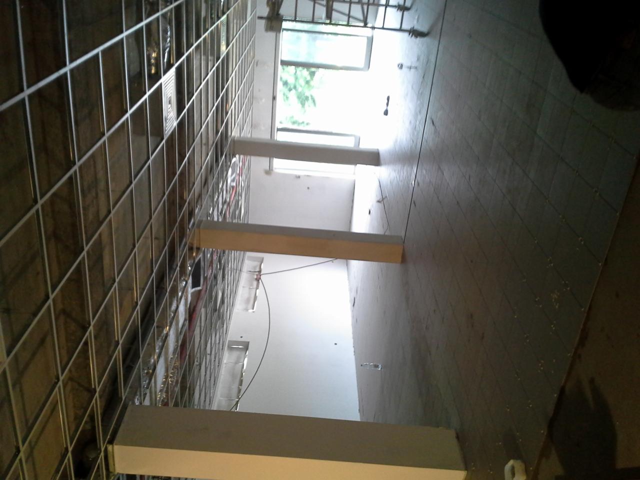 Wnętrze w trakcie prac: metalowa siatka na ścianie, białe słupy i płytki podłogowe. Widok z góry na postęp prac w pomieszczeniu z oknami.