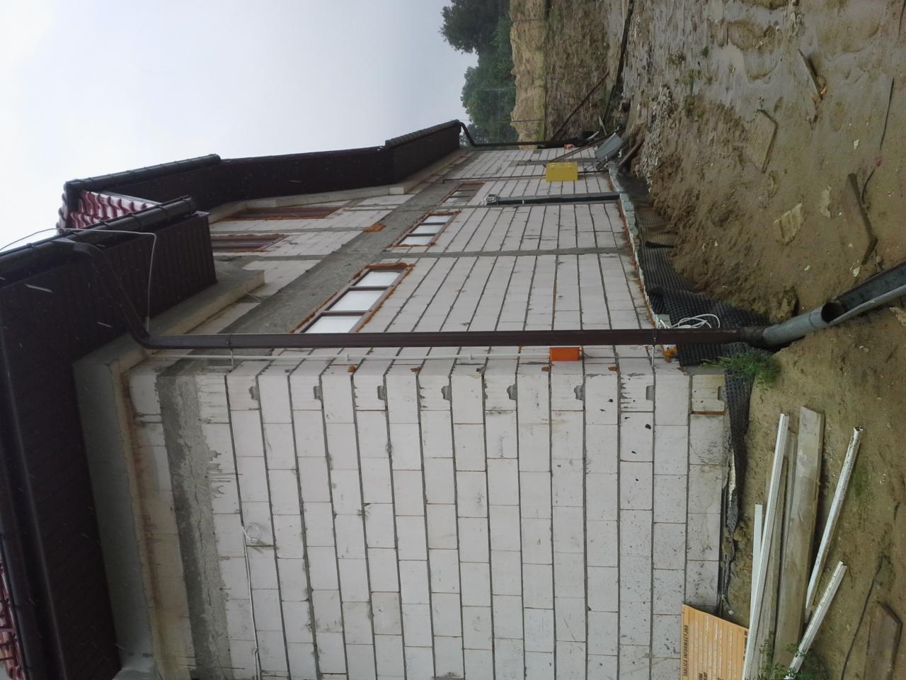 Ściana budynku w trakcie budowy, widać białe bloczki, okna i częściowo zamontowane orynnowanie. Na dole widoczna folia fundamentowa i deski.