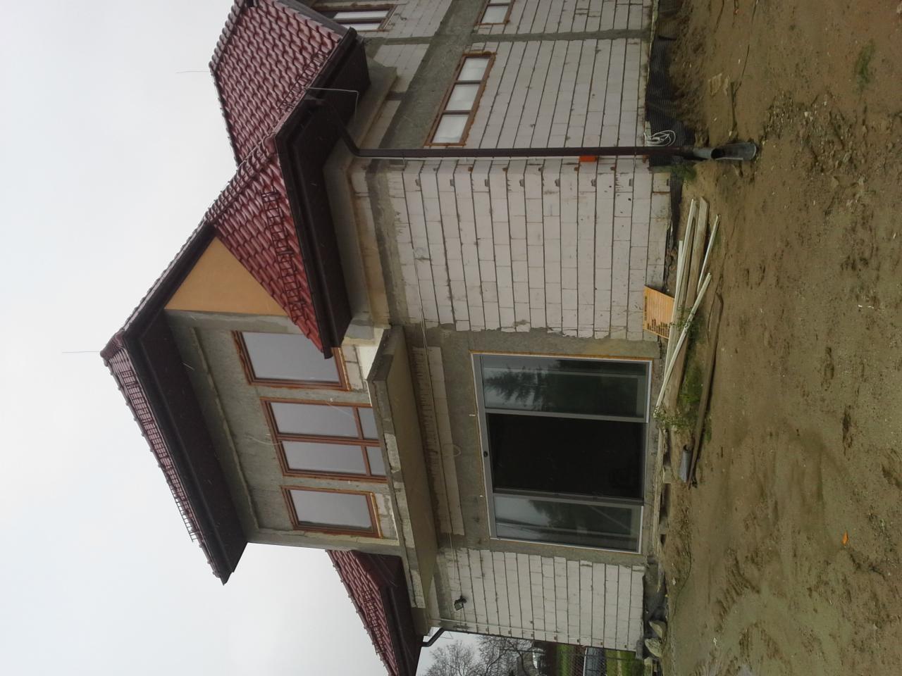 Surowy stan budynku z czerwonym dachem, widoczne okna i białe bloczki. Nieotynkowana elewacja i teren budowy. Ujęcie z dołu, pochmurna pogoda.