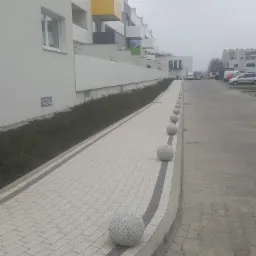 Jasna kostka brukowa ułożona wzdłuż nowoczesnego budynku z kolorowymi balkonami, oddzielona od parkingu rzędem granitowych kul.