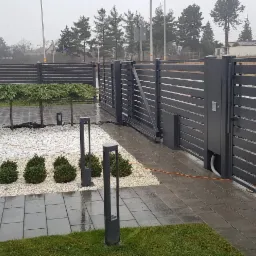 Nowoczesna, pozioma, antracytowa brama wjazdowa przesuwna z automatyką, kostka brukowa, białe kamienie dekoracyjne i nasadzenia w ogrodzie.