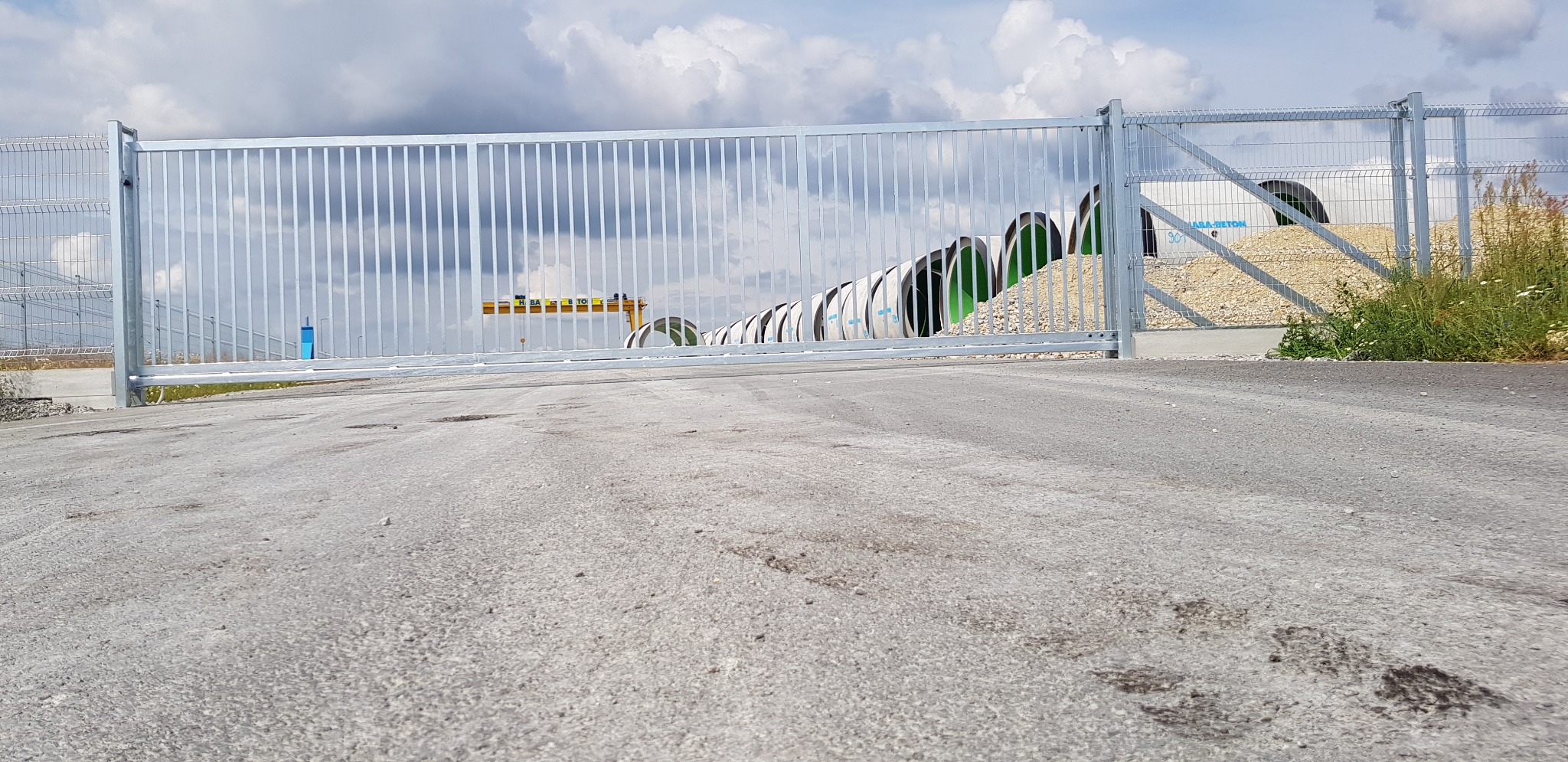 Przesuwna brama wjazdowa z metalowych prętów na tle betonowych rur i żwirowego nasypu, widziana z niskiej perspektywy.
