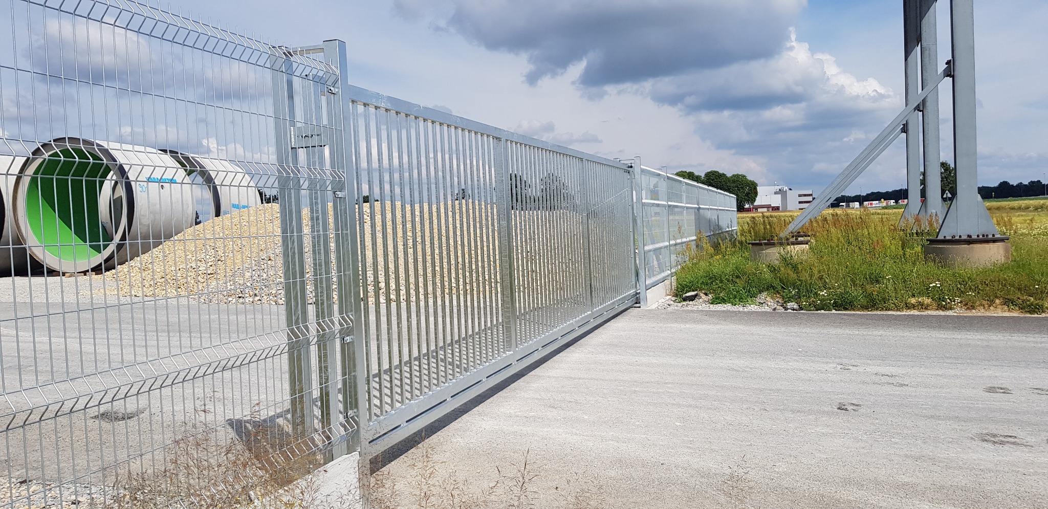 Przesuwna brama przemysłowa z metalową siatką na tle budynków przemysłowych i składowanych rur betonowych.