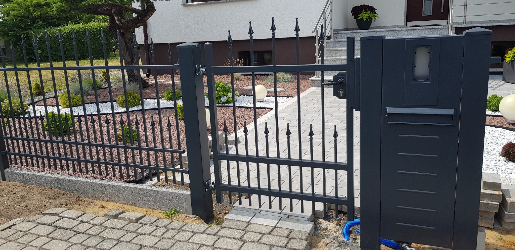 Grafitowa brama wejściowa z furtką i przęsłami ogrodzenia metalowego, widoczne skrzynka na listy i ozdobne groty, w tle ogród z kamieniami ozdobnymi.
