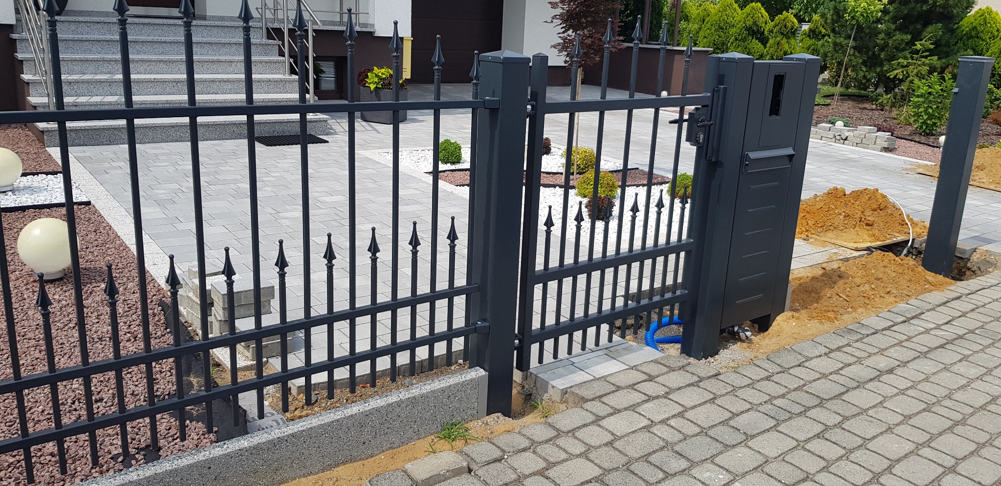 Nowoczesne, grafitowe ogrodzenie metalowe z bramą wjazdową w trakcie montażu, widoczne okablowanie i wykopana ziemia, kostka brukowa na podjeździe.