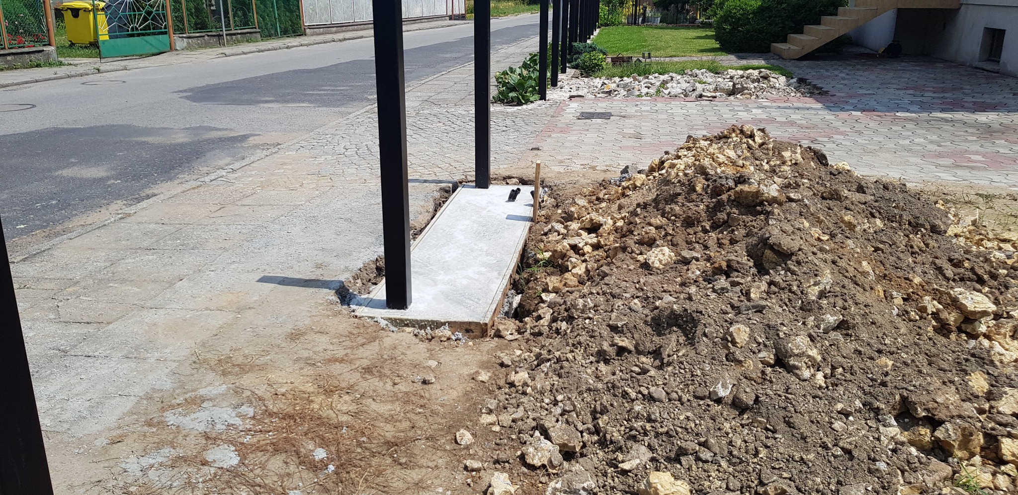 Przygotowanie fundamentu pod czarne słupki ogrodzeniowe, widoczny wykop i pryzma ziemi na tle chodnika i ulicy.