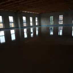 Świeżo wylana, brązowa posadzka w dużym pomieszczeniu z widocznymi oknami i odbiciami światła, zabezpieczona niebieską folią przy ścianach.