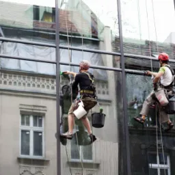Dwóch alpinistów w kaskach myje duże okna budynku z zewnątrz, używając ściągaczek i wisząc na linach; w tle widoczne budynki z cegły.