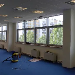 Przestronne biuro z niebieską wykładziną, duże okna z widokiem na budynki i drzewa, odkurzacz przemysłowy na podłodze, przygotowany do czyszczenia.