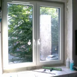 Białe okno PCV, jedna szyba umyta, druga brudna, widok na zielone drzewa i biały samochód, na parapecie leżą akcesoria do mycia okien i butelki z płynem.