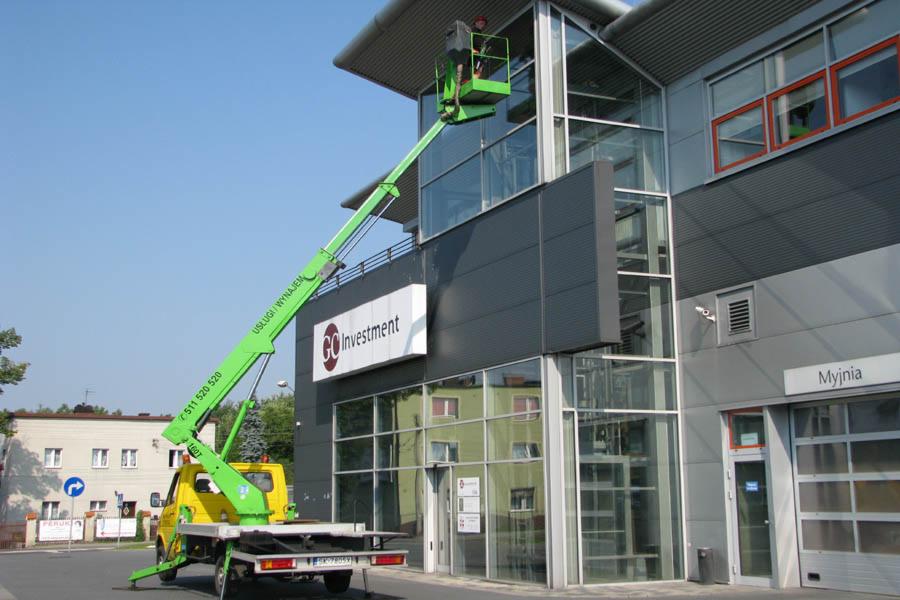 Żółto-zielony podnośnik koszowy przed nowoczesnym budynkiem z przeszkloną fasadą, pracownik w koszu na wysokości czyści okna.