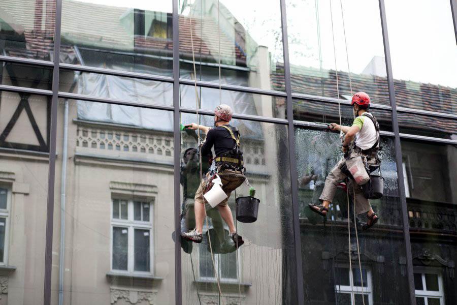 Dwóch alpinistów w kaskach myje duże okna budynku z zewnątrz, używając ściągaczek i wisząc na linach; w tle widoczne budynki z cegły.