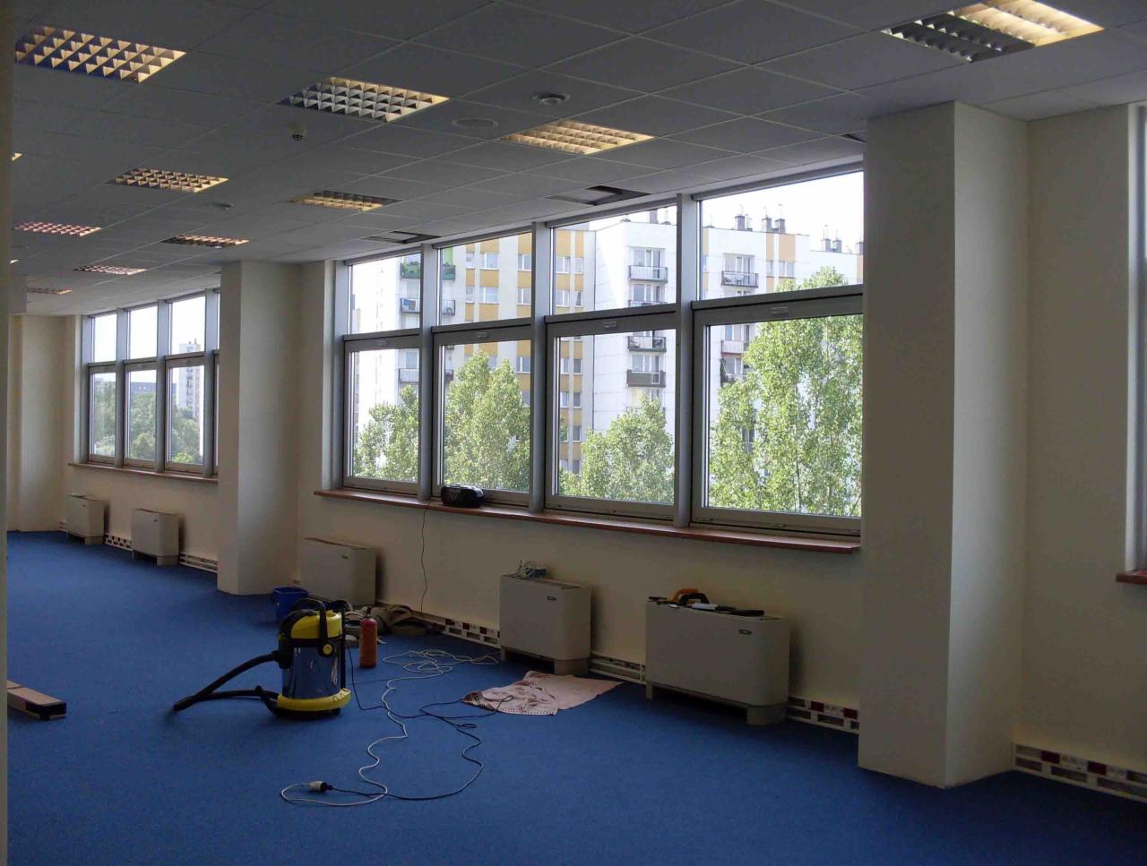 Przestronne biuro z niebieską wykładziną, duże okna z widokiem na budynki i drzewa, odkurzacz przemysłowy na podłodze, przygotowany do czyszczenia.